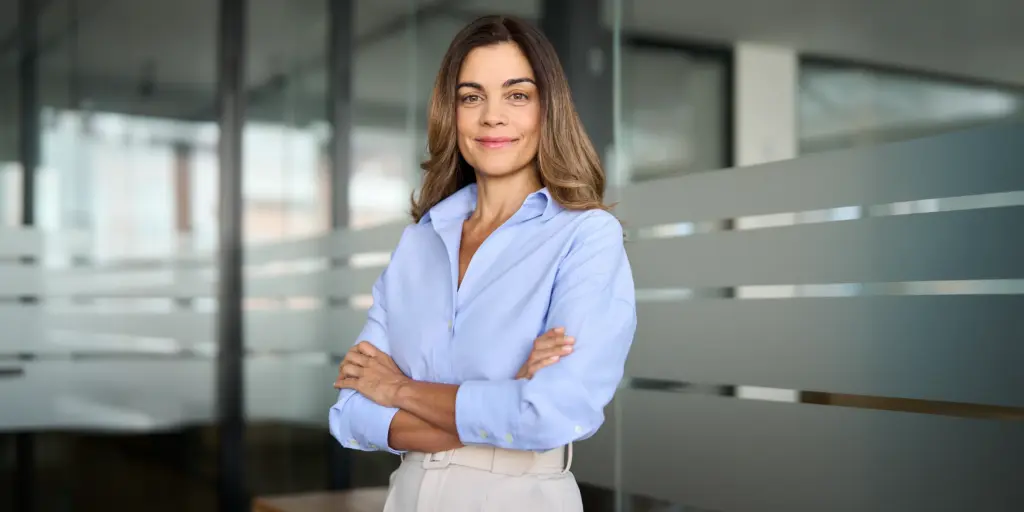 Geschäftsfrau in Büro, trägt hellblaues Hemd und posiert selbstbewusst mit verschränkten Armen vor einer Glaswand.