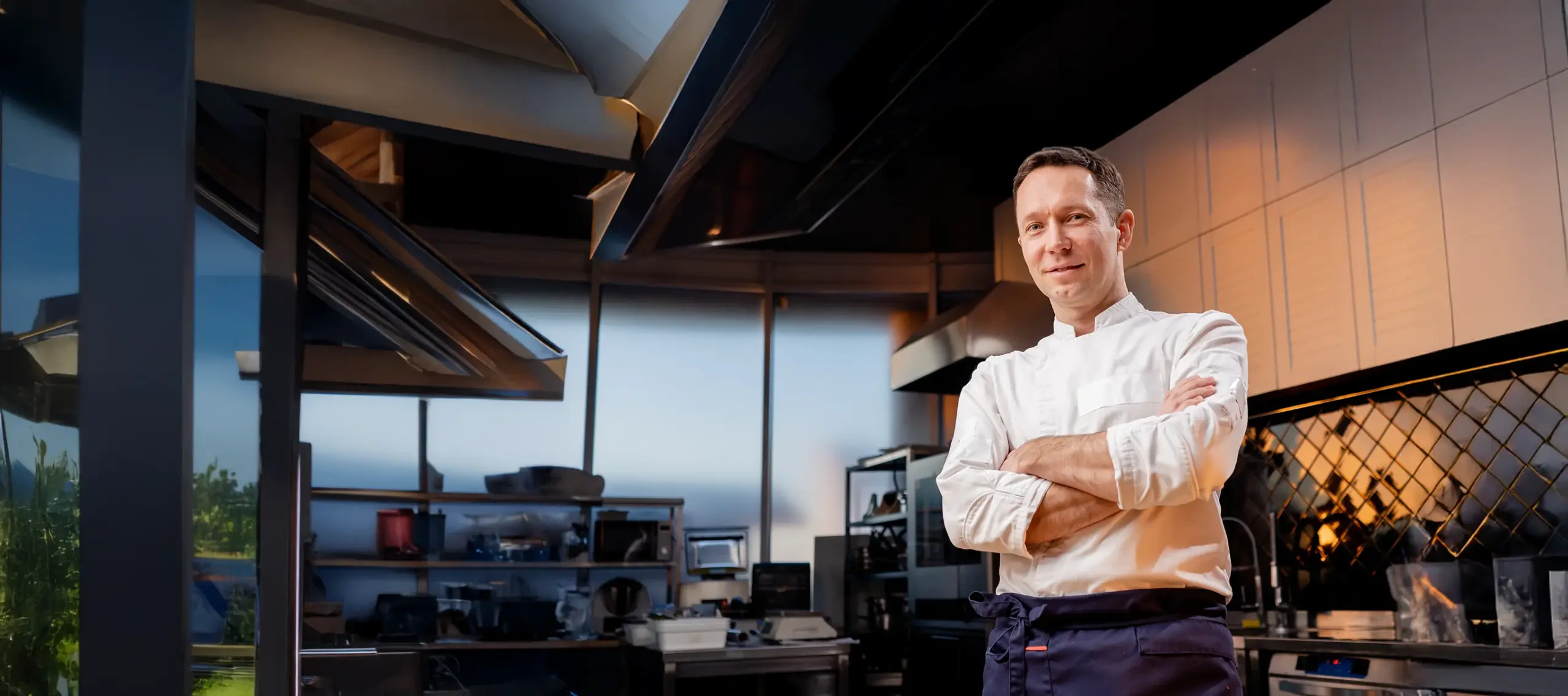 Ein Koch in weißer Uniform steht selbstbewusst in einer modernen Profiküche mit Edelstahlfronten.