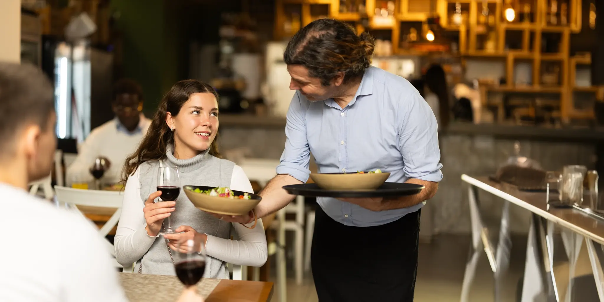Ein Kellner serviert einer Frau im Restaurant ein Essen; beide lächeln. Gemütliche Atmosphäre.